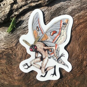 May include: A white sticker with a detailed illustration of a moth with a human-like body. The moth has large wings with orange, white, and gray markings. The moth's body is brown and furry, with red eyes.