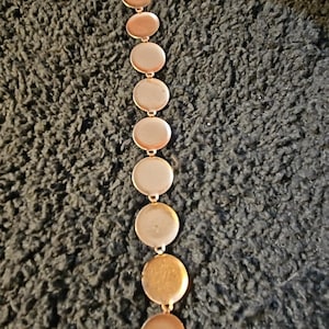 May include: A gold-toned necklace featuring a series of connected, circular pendants. The necklace has a delicate chain and a warm, metallic sheen. The pendants are evenly spaced, creating a flowing, elegant design. The necklace is displayed against a textured, dark background.