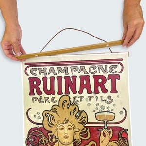 May include: A vintage poster advertising Champagne Ruinart, featuring a woman with flowing blonde hair holding a glass of champagne. The poster has a cream background with red and gold accents. The text "Champagne Ruinart P&egrave;re et Fils" is prominently displayed.