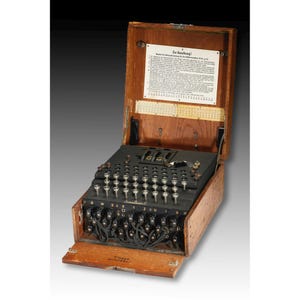 May include: A vintage Enigma encryption machine housed in a wooden case with a hinged lid. The device features a QWERTZ keyboard, a black control panel with numerous metal keys, and a white instructional placard with German text inside the lid.