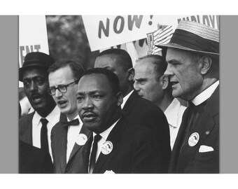 May include: Black and white photograph of a group of men, including Martin Luther King Jr., at a protest. A sign in the background reads "NOW". The men are wearing suits and hats, and some have buttons on their lapels.
