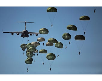 Puede incluir: Un gran avión de transporte militar vuela en un cielo azul despejado con numerosos paracaidistas descendiendo bajo paracaídas de color verde oliva. El avión está situado a la izquierda, mientras los paracaidistas se dispersan por el aire.