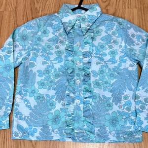 May include: A long-sleeved shirt with a floral pattern in shades of blue and turquoise. The shirt has a ruffled front and a collar. The fabric is covered in a repeating floral design. The shirt is buttoned up the front.