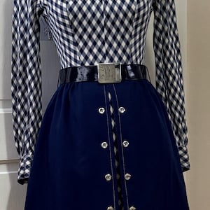 May include: A long-sleeved dress featuring a navy blue and white gingham bodice with a pointed collar. The skirt is solid navy blue with a vertical front opening accented by two parallel rows of silver-toned buttons and a black patent belt.