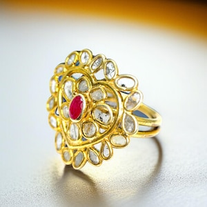 May include: A gold ring with a heart-shaped design. The ring features a central oval ruby surrounded by clear, oval-shaped stones. The ring's band is a simple gold band.