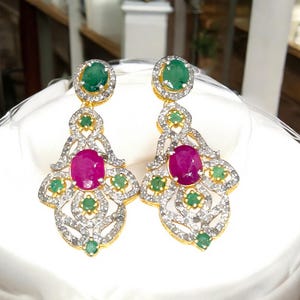 May include: A pair of ornate gold earrings featuring a large oval ruby, surrounded by smaller emeralds and clear gemstones. The earrings have a detailed, openwork design with a green emerald at the top.