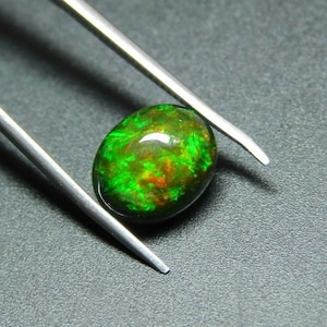 Könnte beinhalten: Ein grüner Opal-Edelstein mit roten und orangefarbenen Blitzen, gehalten von einer Pinzette vor einem dunkelgrauen Hintergrund.