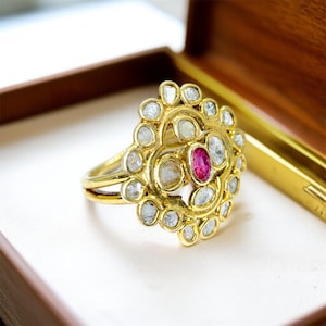 May include: A gold ring with a floral design, featuring a central oval ruby surrounded by numerous smaller, clear gemstones. The ring is set in a gold band and is displayed in a wooden box.
