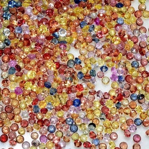 May include: A collection of small, round gemstones in a variety of colors, including yellow, orange, red, pink, blue, and purple. The gemstones are scattered across a white surface, creating a colorful and sparkling display. These gemstones could be used for jewelry making.