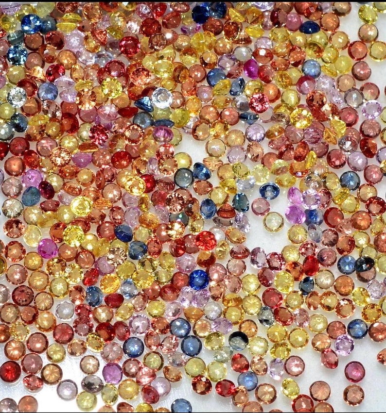 May include: A collection of small, round gemstones in a variety of colors, including yellow, orange, red, pink, blue, and purple. The gemstones are scattered across a white surface, creating a colorful and sparkling display. These gemstones could be used for jewelry making.