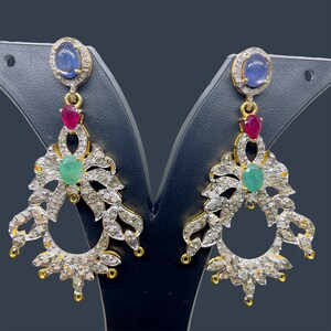 Puede incluir: Un par de pendientes ornamentados con una piedra preciosa ovalada azul, una piedra preciosa roja y una piedra preciosa verde. Los pendientes tienen un diseño floral con pequeñas gemas claras y detalles dorados. Se exhiben en un soporte negro.