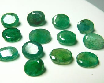 May include: A collection of oval-cut emerald gemstones in varying shades of green. The gemstones are faceted, displaying multiple reflective surfaces. The arrangement is on a white surface, highlighting the vibrant color and clarity of the emeralds.