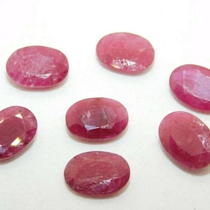 Może przedstawiać: Siedem owalnych rubinów o różnych rozmiarach jest prezentowanych na białym tle. Kamienie szlachetne wykazują głęboki, bogaty czerwony odcień z widocznymi wewnętrznymi teksturami i wtrąceniami. Fasetki wychwytują światło, tworząc efekt iskrzenia. Rubiny te nadają się do wyrobu biżuterii.