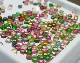 May include: A collection of round, faceted gemstones in various shades of green, pink, and blue. The gemstones are arranged in a white tray.