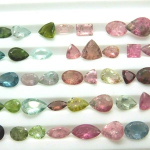 Könnte beinhalten: Sortiment an losen Edelsteinen in verschiedenen Farben, darunter Grün, Rosa, Blau und Braun. Die Edelsteine ​​werden auf einer weißen Oberfläche präsentiert.