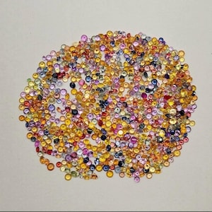 May include: A collection of small, round gemstones in a variety of colors, including yellow, orange, pink, purple, and blue. The gemstones are scattered across a white surface, creating a colorful and textured display. The gemstones appear to be polished and faceted.