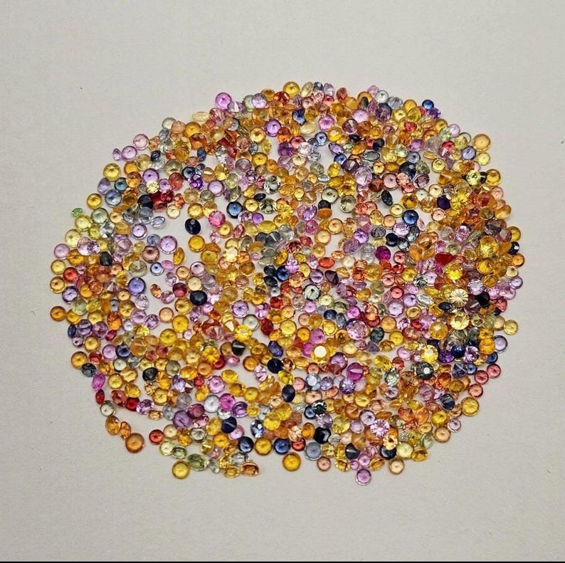 May include: A collection of small, round gemstones in a variety of colors, including yellow, orange, pink, purple, and blue. The gemstones are scattered across a white surface, creating a colorful and textured display. The gemstones appear to be polished and faceted.