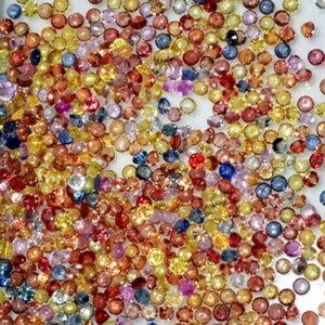 May include: A collection of small, round gemstones in various colors, including red, yellow, blue, and purple. The gemstones are scattered across a white surface, creating a colorful and sparkling display. The gemstones appear to be faceted, reflecting light.