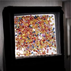 May include: A collection of colorful gemstones, including red, yellow, blue, and orange, are displayed in a square, illuminated frame. The gems are small and round, creating a vibrant, textured surface. The frame is dark, contrasting with the bright gems.