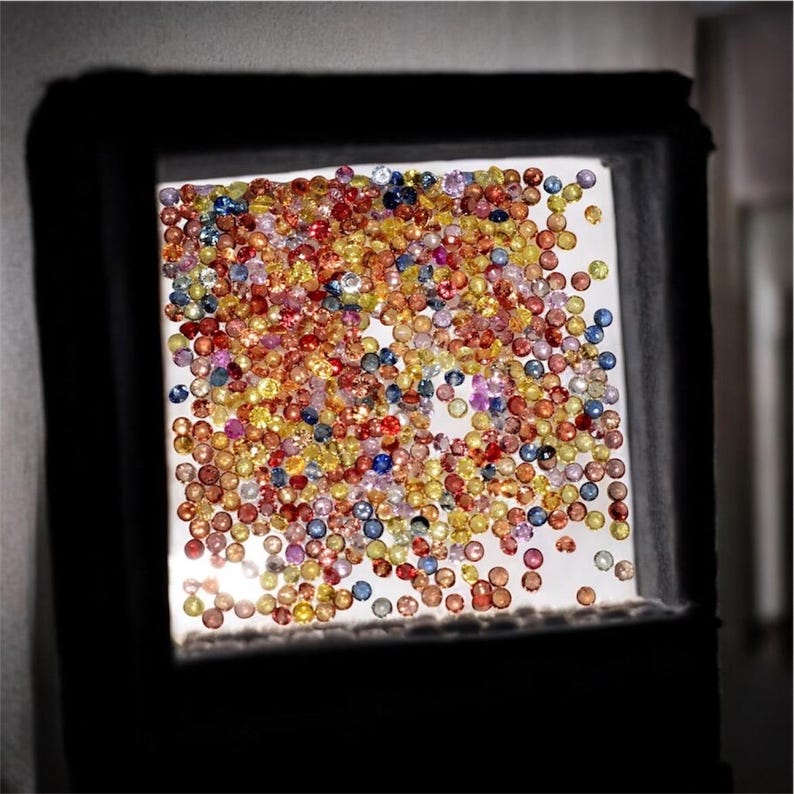 May include: A collection of colorful gemstones, including red, yellow, blue, and orange, are displayed in a square, illuminated frame. The gems are small and round, creating a vibrant, textured surface. The frame is dark, contrasting with the bright gems.