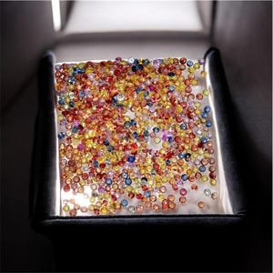 May include: A collection of colorful gemstones, including red, yellow, blue, and orange, are displayed on a white surface. The gems are small and round, filling the frame. The image is lit from above, creating shadows.