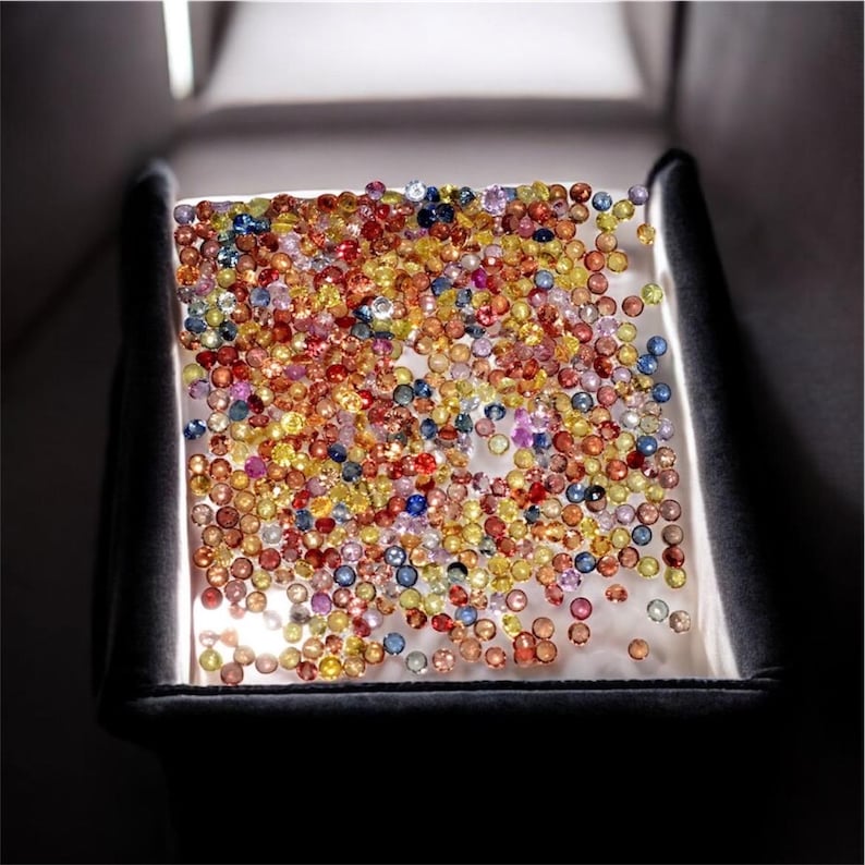 May include: A collection of colorful gemstones, including red, yellow, blue, and orange, are displayed on a white surface. The gems are small and round, filling the frame. The image is lit from above, creating shadows.