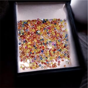 May include: A collection of small, colorful gemstones in a display box. The gems are a mix of red, yellow, blue, and orange hues, creating a vibrant and eye-catching display. The box is dark, contrasting with the bright gems.
