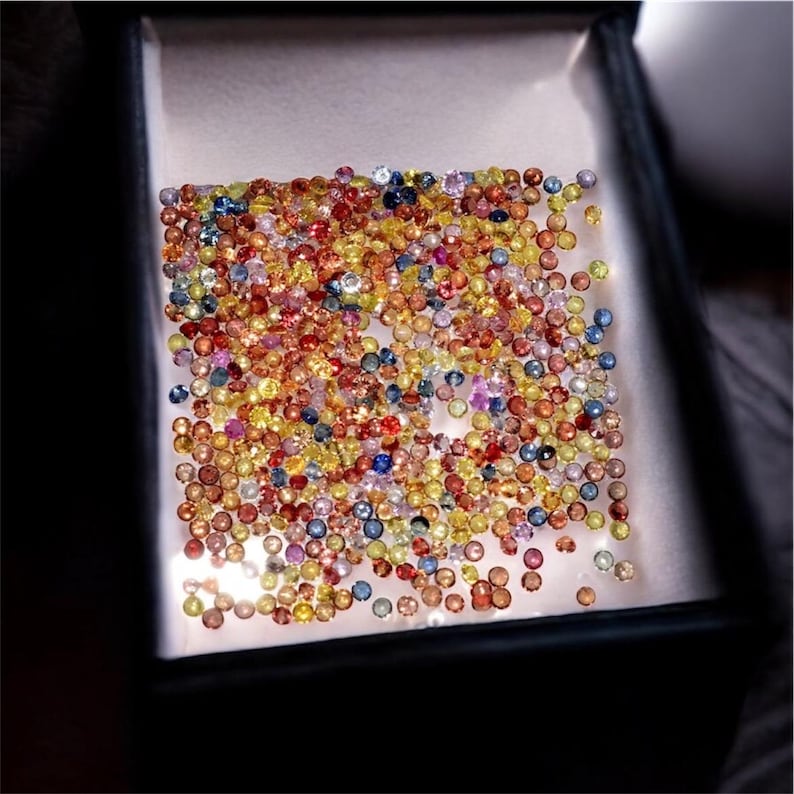 May include: A collection of small, colorful gemstones in a display box. The gems are a mix of red, yellow, blue, and orange hues, creating a vibrant and eye-catching display. The box is dark, contrasting with the bright gems.