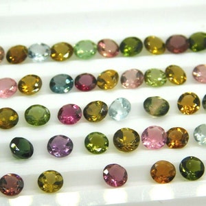 May include: A collection of round, faceted gemstones in various colours, including green, pink, blue, and brown. The gemstones are arranged in rows on a white surface.