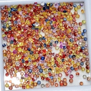 May include: A collection of small, round gemstones in a variety of colors, including red, orange, yellow, pink, blue, and purple. The gemstones are scattered across a white surface, creating a colorful and sparkling display.
