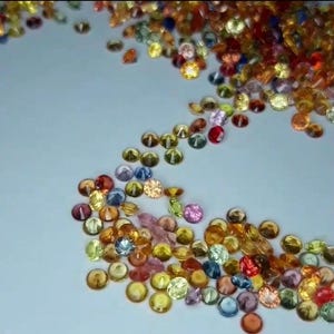 May include: A scattering of small, colorful gemstones in various shades of yellow, orange, pink, blue, and red. The gemstones are round and faceted, with a slight dome shape. The gems are scattered on a light blue surface.