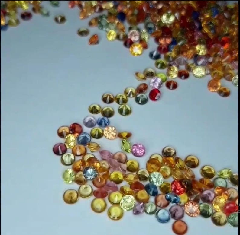 May include: A scattering of small, colorful gemstones in various shades of yellow, orange, pink, blue, and red. The gemstones are round and faceted, with a slight dome shape. The gems are scattered on a light blue surface.