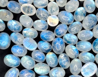 May include: A collection of oval-shaped moonstone cabochons. The stones display a milky white base with iridescent blue flashes. The polished gemstones are scattered across a dark surface, showcasing their natural beauty and potential for jewelry making.