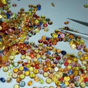 May include: A collection of small, round gemstones in various colors, including yellow, orange, pink, blue, and green. A pair of tweezers is visible, suggesting the gems are being handled or sorted. The gems are scattered on a light-colored surface.