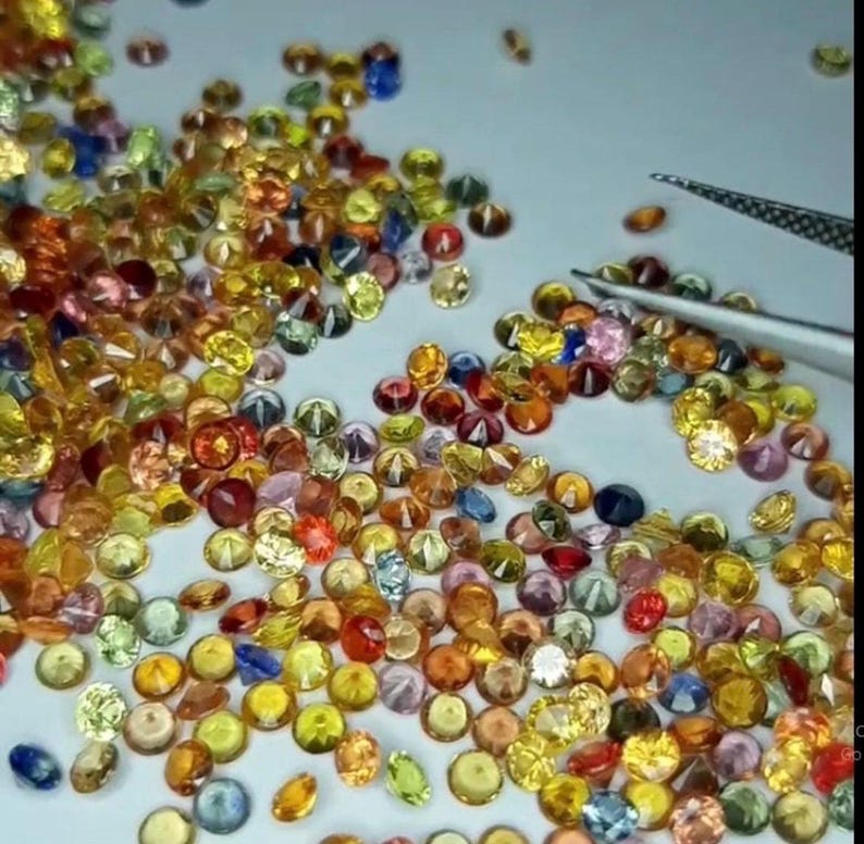 May include: A collection of small, round gemstones in various colors, including yellow, orange, pink, blue, and green. A pair of tweezers is visible, suggesting the gems are being handled or sorted. The gems are scattered on a light-colored surface.