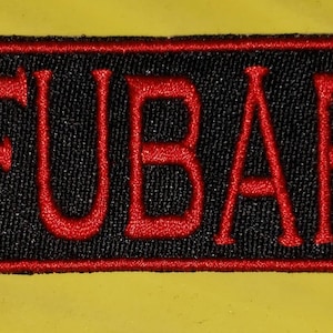 May include: A rectangular black patch with the word "FUBAR" embroidered in red. The patch has a red border and is set against a yellow background. The letters are in a bold, sans-serif font.