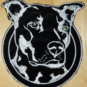 May include: Embroidered patch featuring a black dog's head with green eyes, outlined in white. The dog is centered within a black circle, also outlined in white. The patch is on a wooden surface.