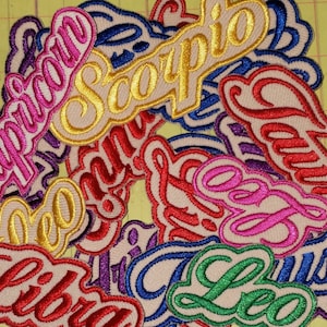 May include: A collection of embroidered patches featuring various zodiac signs in cursive script. The patches have a cream-colored background with colorful stitching in shades of pink, gold, red, blue, and green. The patches are in a variety of shapes.