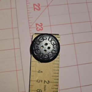 May include: A black button with the word "UGG" embossed around the edge, featuring four holes for sewing. The button is approximately 1.5 inches in diameter and is placed on a wooden ruler.