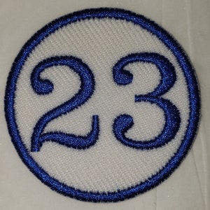 May include: Circular white patch with the number 23 embroidered in blue. The number is inside a blue embroidered circle. The patch has a textured, woven appearance.