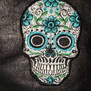 May include: A white sugar skull embroidered patch with teal and green floral details. The skull has black eyes and a toothy grin.
