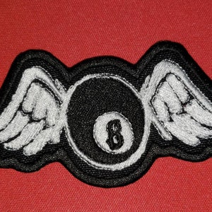 May include: Embroidered patch featuring a black eight ball with white wings. The patch has a black border and is set against a red background. The number 8 is visible on the ball.