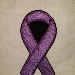 May include: Embroidered purple awareness ribbon with a black center, set against a textured, off-white background. The ribbon has a detailed, stitched appearance, with a dark purple outline.