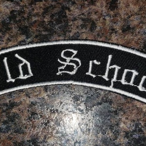 May include: Black, arched patch with white embroidered text reading "Old School." The patch has a white border and is likely designed to be sewn onto clothing or accessories. The patch is on a brown and black speckled surface.