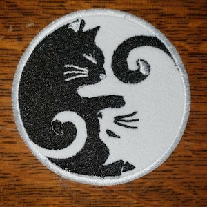 May include: An oval patch featuring a Yin and Yang design with cats. One cat is black, the other white, with whiskers and a swirling tail. The patch has a white border with silver stitching, set against a wooden background.