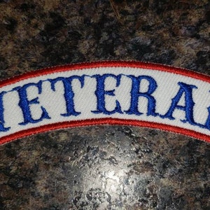 May include: A white, arched patch with the word "VETERAN" embroidered in blue capital letters. The patch has a red border and is designed to be sewn onto clothing or other fabric items.