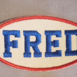 May include: White oval patch with a red border. The word "FRED" is embroidered in blue.