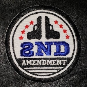 May include: Circular patch with a black border, featuring two black gun silhouettes, red star accents, and the text "2ND AMENDMENT" in blue and white. The patch is on a black leather-like surface.