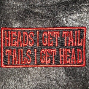 May include: Embroidered patch with the text "HEADS I GET TAIL, TAILS I GET HEAD" in red thread on a black background, framed by a red border. The patch is set against a textured black surface.