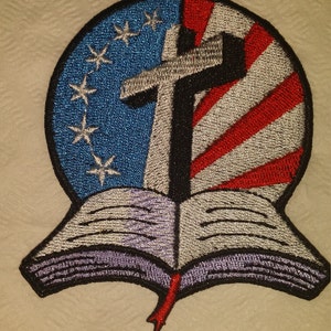 May include: An embroidered patch featuring a cross over an American flag with a bible open underneath. The cross is black, the flag is red, white, and blue, and the bible is white with purple accents.
